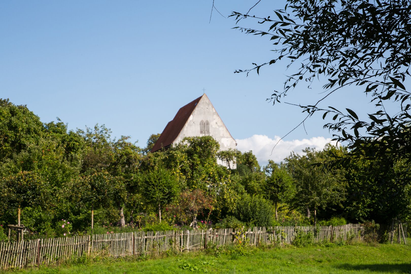 Altes Land-Kirchengebäude in einem Obstbaumgarten. Davor ein Holzzaun. Im Vordergrun eine Wiese.