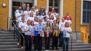Eine Menschengruppe mit weißen T-shirts steht vor dem eingang eines Hauses (Vineta Bürgerhaus). die erste Reihe hält eine Kette aus kleinen Flaggen.