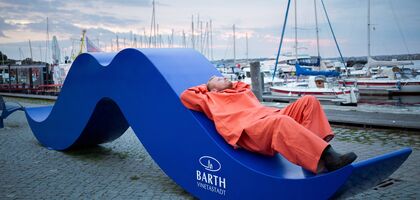 Ein Fischer in Ölzeug liegt gemütlich auf einem riesigen blauen Bart am Hafen