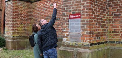 eine Frau und ein Mann stehen vor einem Backsteingebäude. vor ihnen eine Hinweistafel. Er zeigt mit dem Finger am ausgestreckten Arm nach oben uns zeigt der Frau etwas.