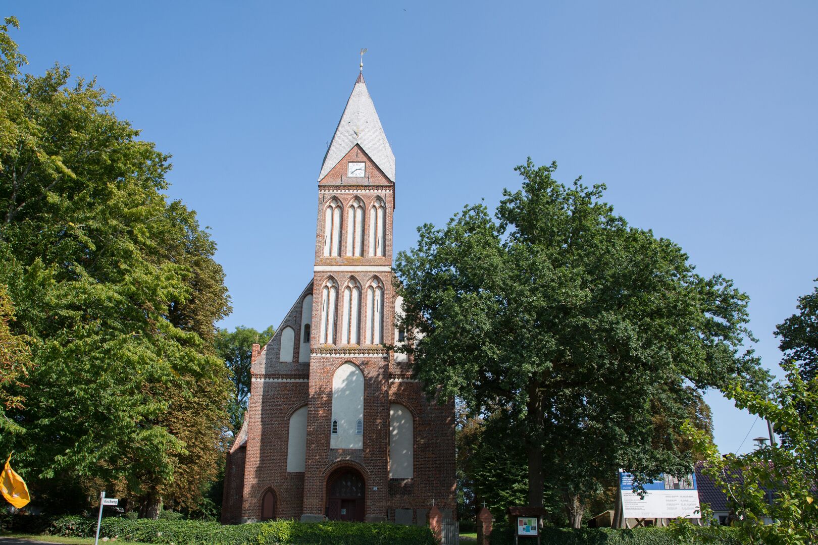 Kirche mit schmalem Kirchturm an den Giebel erbaut inmitten von Bäumen und Hecken
