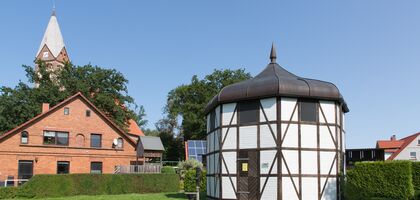 Achteckiges Häuschen Fachwerk weiß vermauert, im Hintergrund die Kirche